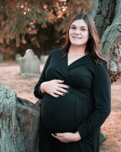 A smiling pregnant woman in a flowing black dress poses against an ancient tree in a Salem, Massachusetts, graveyard, with historic headstones softly visible in the background.