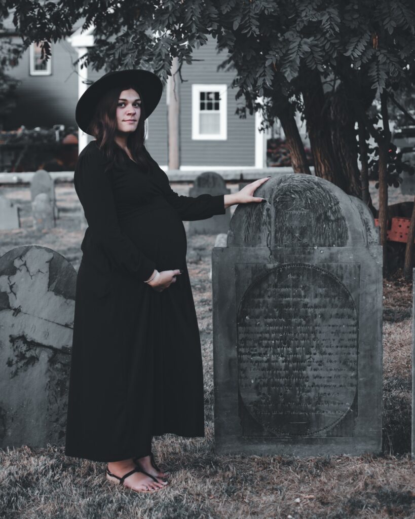 A pregnant woman dressed in black stands beside an aged gravestone in a historic burial ground in Salem, Massachusetts, surrounded by trees and weathered tombstones.