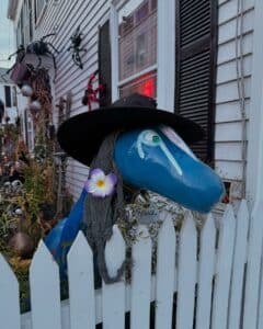 A blue creature statue named Reggie in Salem Massachusetts is dressed up with a witch hat, gray scarf, and a purple flower. Halloween decorations, including large spiders and skulls, cover the house behind the white picket fence.