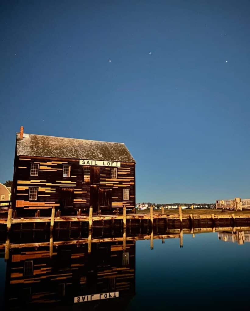 The historic Sail Loft in Salem, Massachusetts, is reflected in the still waters at night under a starry sky. The dark wooden building features light patches and a simple white sign.