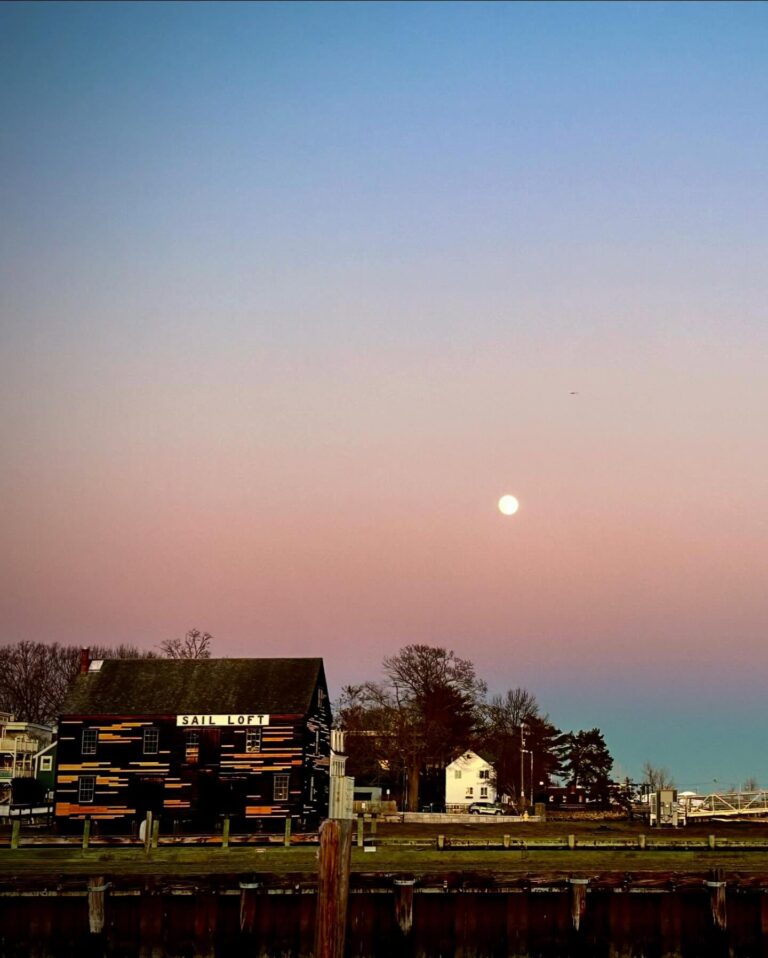 The Sail Loft in Salem, Massachusetts, sits by the water during dusk, with a pastel pink and blue sky and a full moon rising in the background. The dark wooden building reflects in the calm harbor.