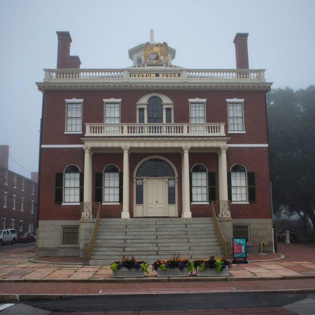 The historic Salem Custom House in Salem, Massachusetts, with its grand brick façade, white columns, and gold eagle, surrounded by a misty fog and vibrant flowerbeds.