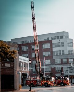 Salem Fire Department ladder truck extending its towering ladder in front of a red brick fire station, with additional fire trucks parked nearby and the cityscape of Salem, Massachusetts, in the background.