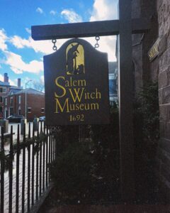 The Salem Witch Museum sign in Salem, Massachusetts, features a dark wooden design with gold lettering and an image of a witch and child, standing near the stone exterior of the museum.