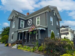 An artistic depiction of the Satanic Temple in Salem, Massachusetts, featuring a dark gray historic house with a porch, a pentagram emblem, and a garden framed by intricate wrought-iron gates.