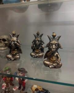A trio of Baphomet-inspired "See No Evil, Hear No Evil, Speak No Evil" figurines displayed on a glass shelf in Salem, Massachusetts, alongside other occult-themed items.
