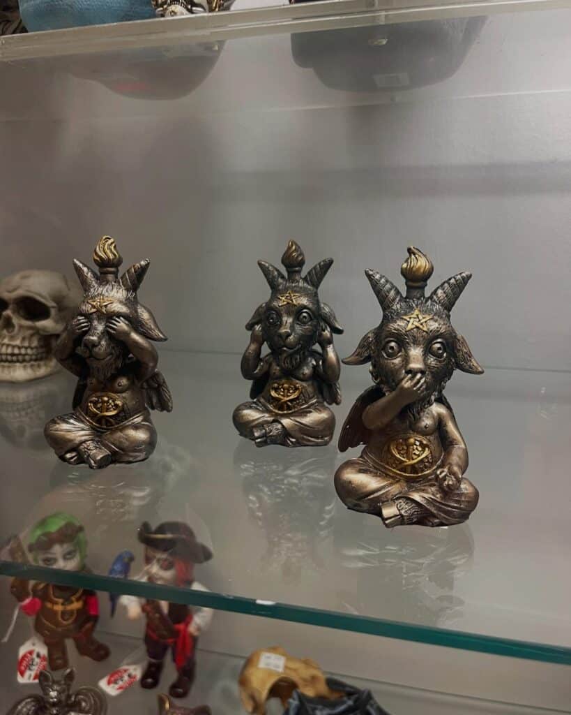  A trio of Baphomet-inspired "See No Evil, Hear No Evil, Speak No Evil" figurines displayed on a glass shelf in Salem, Massachusetts, alongside other occult-themed items.