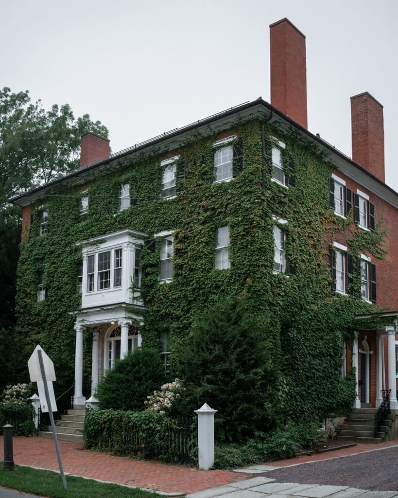 A charming ivy-covered historic mansion in Salem, Massachusetts, featuring red brick chimneys, a classic white entrance, and lush greenery surrounding the property.