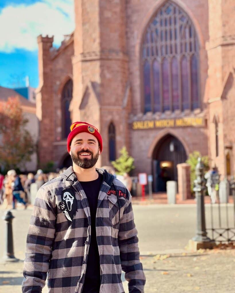 A man wearing a red beanie and a black-and-white flannel stands smiling in front of the Salem Witch Museum in Salem, Massachusetts. The museum's gothic stone facade and arched windows are visible in the background.