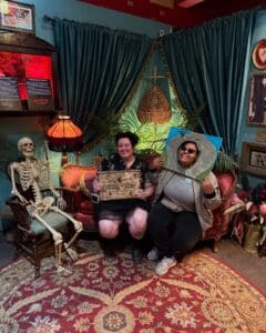 Two visitors seated inside the Salem Witch Board Museum in Salem, Massachusetts. One holds a vintage Ouija board and smiles, while the other poses with a creative costume piece resembling a painted stone. A skeleton sits to the left on a chair, adding to the museum's mystical decor with its velvet drapes, vintage lamp, and ornate furniture.