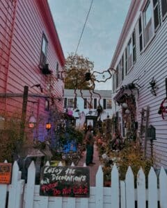 A narrow alley in Salem Massachusetts is decorated with Halloween decorations including large spiders, skeletons, and glowing lights. A sign on the white picket fence reads "Enjoy the decorations, please do not enter."