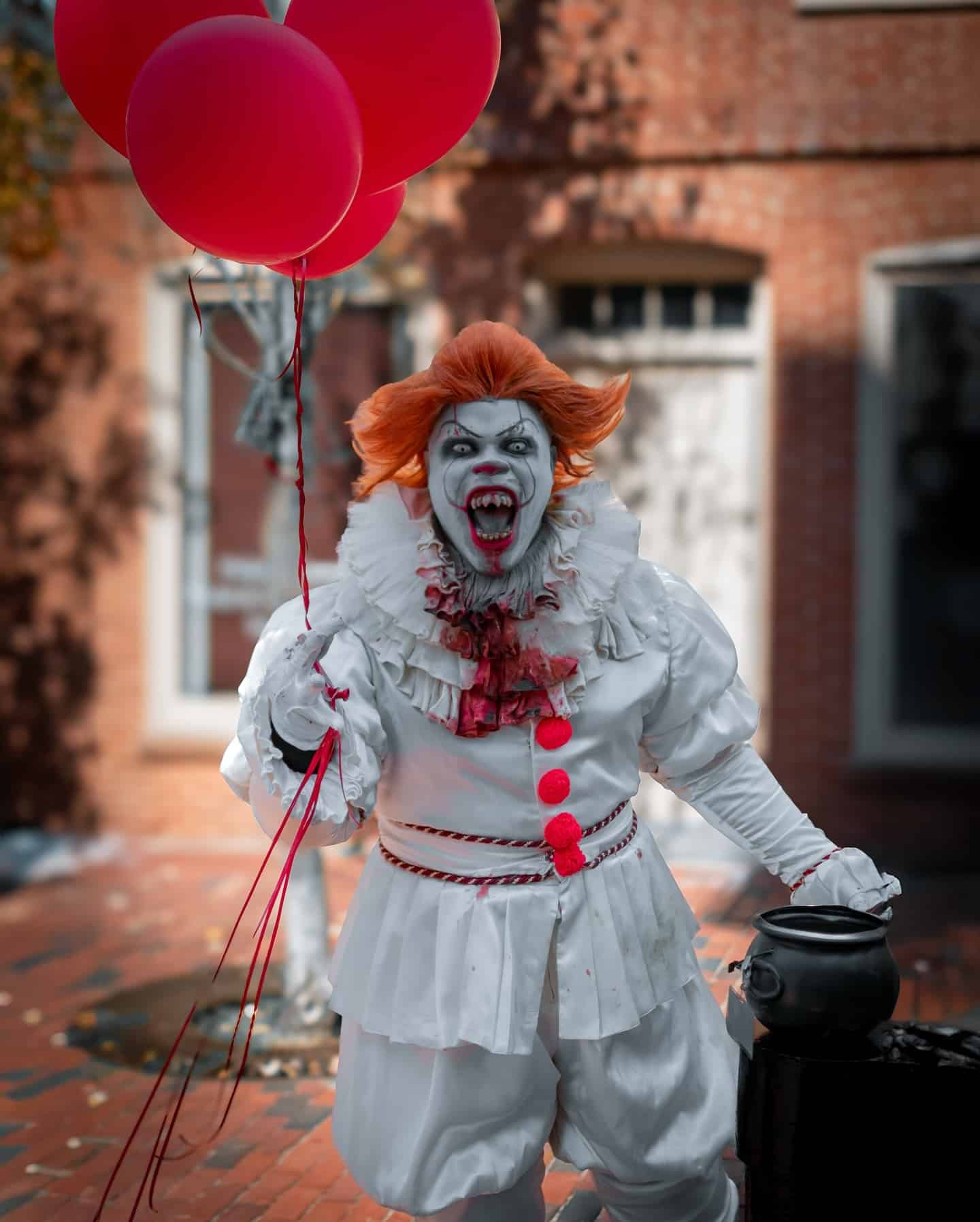A detailed Pennywise cosplay in Salem, featuring the iconic clown holding red balloons and wearing a blood-stained ruffled costume on a brick street backdrop.