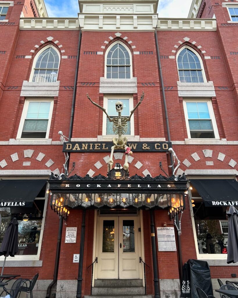 The entrance of Rockafellas in Salem, Massachusetts, featuring a skeleton decoration perched on the historic red-brick Daniel Low & Co. building.
