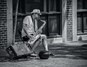 A black-and-white image of a street musician in Salem, Massachusetts, playing a saxophone while seated on a folding chair, with a decorated case labeled "Joe's Sax Appeal" and a small collection pot nearby.