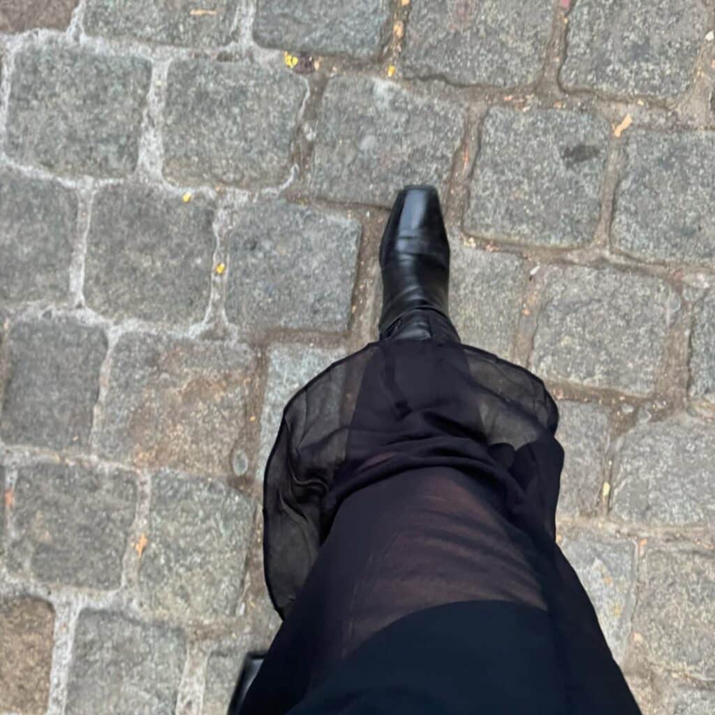 A person wearing a sheer black skirt and black pointed boots walking on a cobblestone street in Salem, Massachusetts, showcasing a fashionable and moody look.