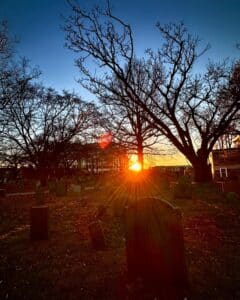 The sun sets over the Old Burying Point Cemetery in Salem, Massachusetts, casting long shadows over gravestones and bare trees. The golden light filters through the branches, creating a peaceful yet eerie atmosphere.