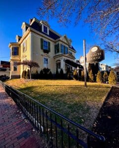 The Coach House in Salem, Massachusetts, a beautifully restored yellow Victorian building with green shutters, sits on a well-manicured lawn under a bright blue sky. A sign in front reads "The Coach House, Est. 1879."