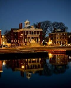 The historic Custom House in Salem, Massachusetts, is illuminated at night and reflected in the still waters of the harbor. The red brick building stands against a deep blue sky with warm lights glowing from nearby structures.