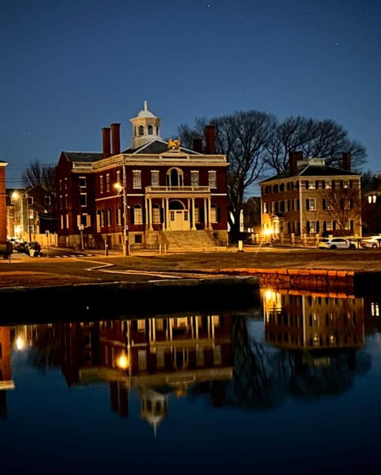 The historic Custom House in Salem, Massachusetts, is illuminated at night and reflected in the still waters of the harbor. The red brick building stands against a deep blue sky with warm lights glowing from nearby structures.