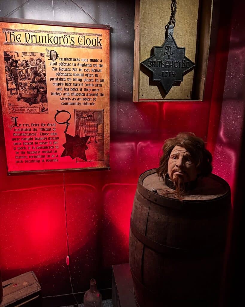 A display at Salem's Museum of Torture featuring a barrel punishment called the Drunkard's Cloak, with a mannequin's head emerging from a barrel and an informational sign detailing its use in historical punishment.