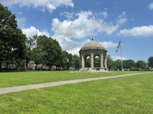 The iconic gazebo at Salem Common in Salem, Massachusetts, surrounded by lush green lawns, mature trees, and a clear blue sky with an American flag waving nearby.