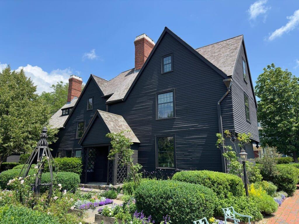 The House of the Seven Gables in Salem, Massachusetts, with its classic dark wood exterior, steep rooflines, and a lush garden filled with colorful flowers and greenery under a bright blue sky.