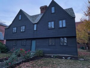 The John Ward House in Salem, Massachusetts, photographed at dusk with its dark wooden exterior and leaded glass windows, framed by a small garden and red roses.