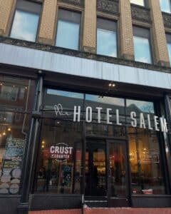 The front entrance of The Hotel Salem in Salem, Massachusetts, features large glass windows and doors. A sign for Crust at The Counter, a restaurant inside, is displayed next to the entrance.