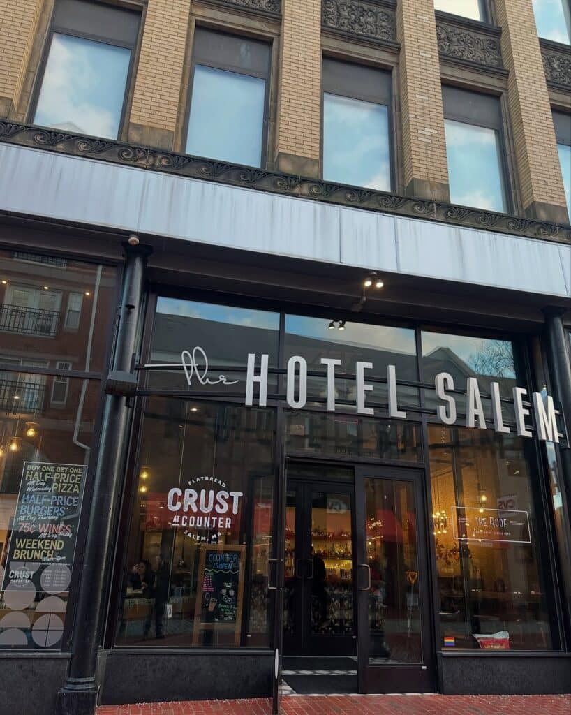 The front entrance of The Hotel Salem in Salem, Massachusetts, features large glass windows and doors. A sign for Crust at The Counter, a restaurant inside, is displayed next to the entrance.