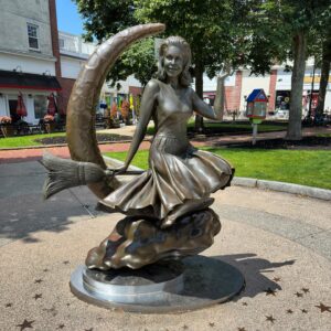 The famous Bewitched statue in Salem, Massachusetts, featuring Samantha Stephens sitting on a crescent moon with her broom, surrounded by a bright park setting and a clear blue sky.