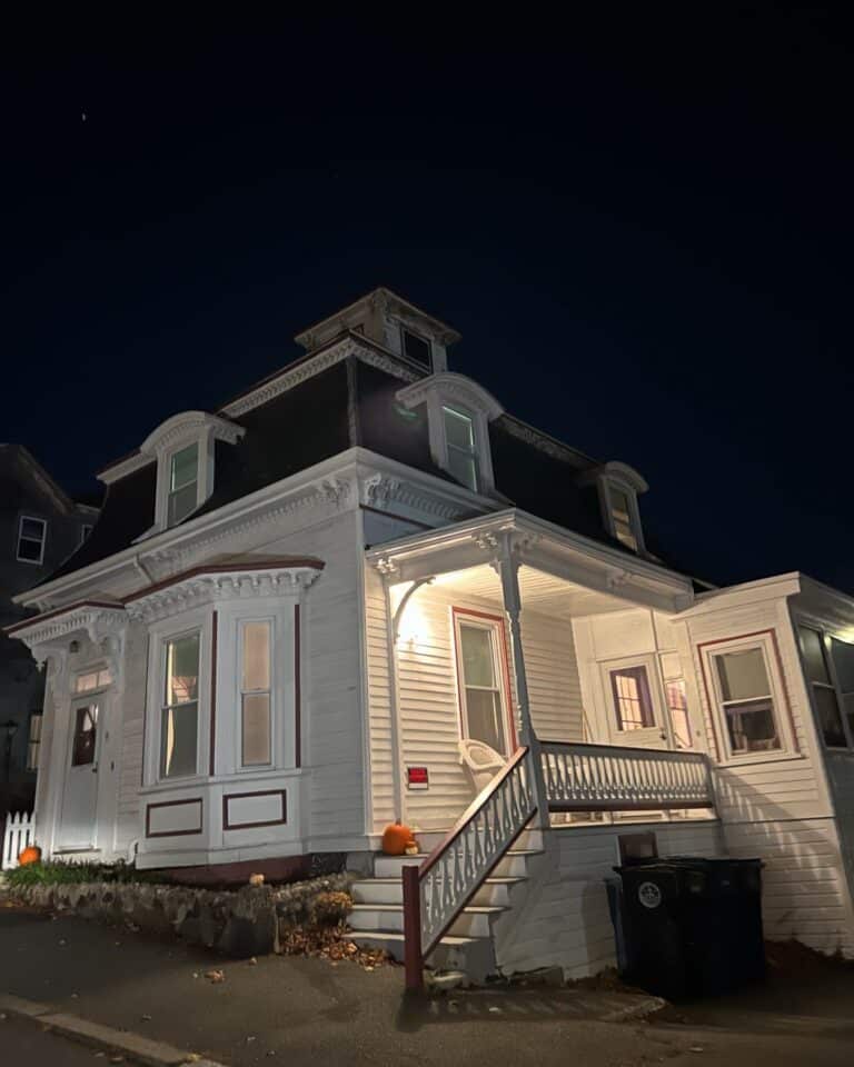 The famous Hocus Pocus House in Salem, Massachusetts, stands illuminated at night, with a white Victorian-style exterior, ornate trim, and a cozy front porch. Pumpkins sit on the steps, adding a Halloween touch.