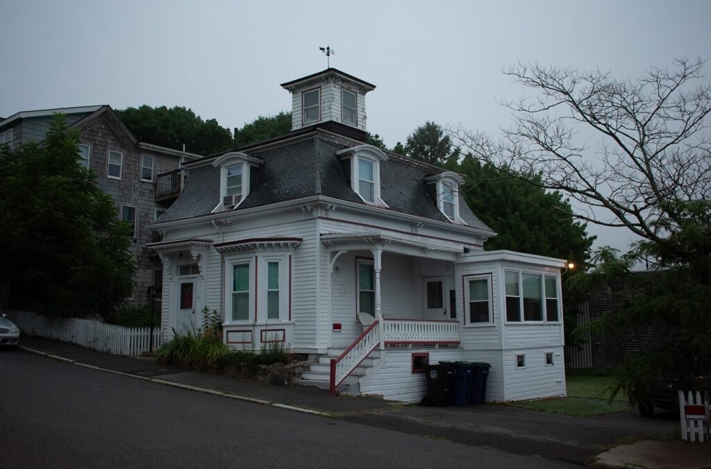The Hocus Pocus house in Salem, Massachusetts, shown in a calm evening setting with its distinctive mansard roof and cupola, surrounded by lush greenery and a quiet street.