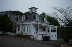 The Hocus Pocus house in Salem, Massachusetts, shown in a calm evening setting with its distinctive mansard roof and cupola, surrounded by lush greenery and a quiet street.