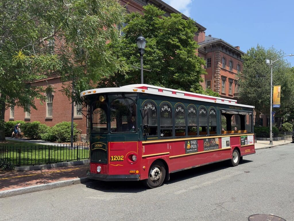 The Salem Trolley in Salem, Massachusetts, parked on a tree-lined street near historic red brick buildings on a sunny day.