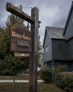 A wooden sign reading "Witch House" hangs from a weathered wooden post in front of the historic Witch House in Salem, Massachusetts, with the dark gray building and overcast skies creating a mysterious atmosphere.