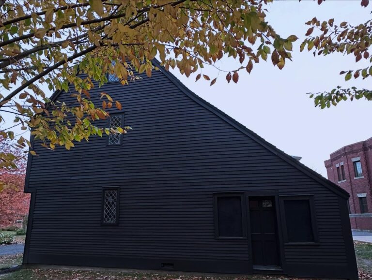 A side view of the John Ward House in Salem, Massachusetts, with its dark wooden siding, sloped roof, and diamond-shaped windows, framed by golden autumn leaves.