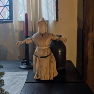 A handcrafted corn husk poppet displayed in a historic Salem home, set on a wooden table with a red candle, ceramic jug, and light streaming through a leaded glass window.