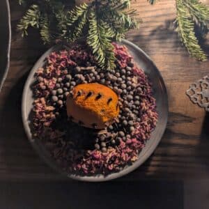 A clove-studded orange sits on a plate surrounded by dried rose petals and spices, with evergreen branches in the background, creating a warm and festive scene in Salem, Massachusetts.