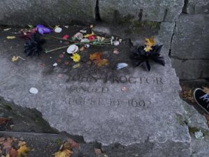 John Proctor’s memorial stone at the Salem Witch Trials Memorial in Salem, Massachusetts, adorned with flowers, coins, and tokens of remembrance.