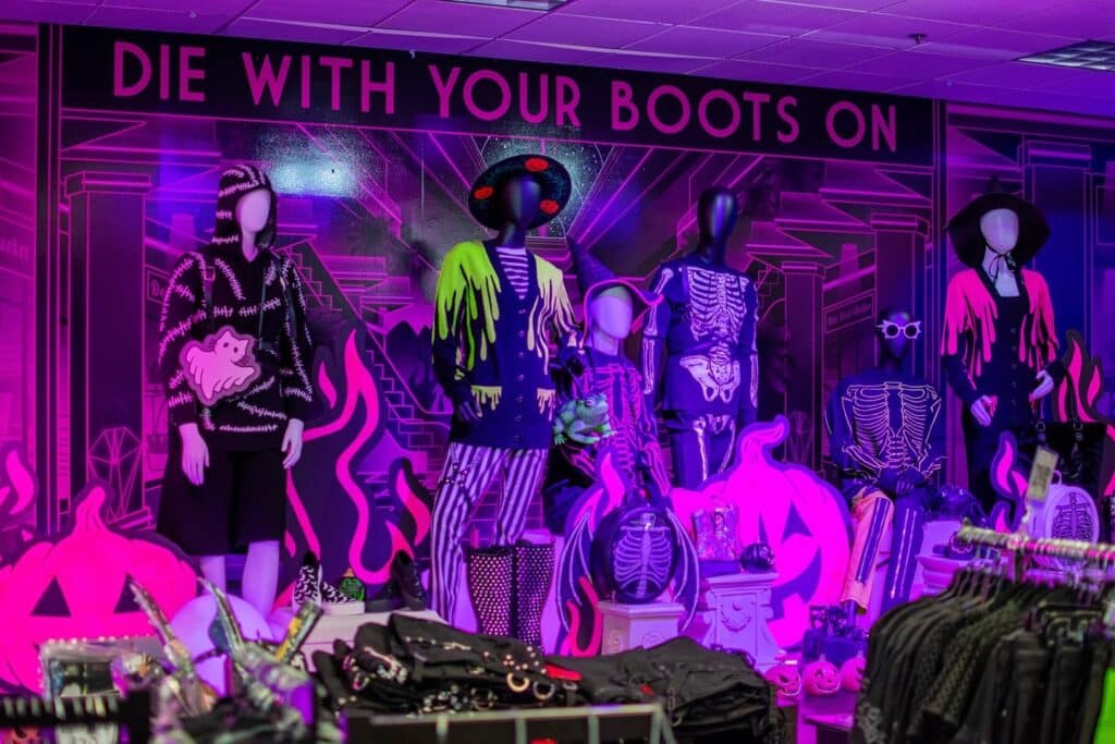 A neon-lit Halloween fashion display in Salem, featuring mannequins dressed in skeleton outfits, spooky sweaters, and witch hats with glowing pumpkins and pink decor.