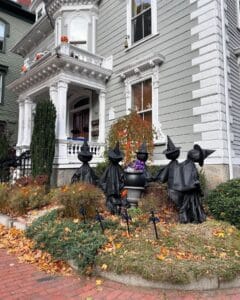 A Victorian-style home in Salem, Massachusetts, decorated for Halloween with a group of witch-like figures in black cloaks and hats gathered in the yard. The house features ornate white trim, jack-o'-lanterns on the balcony, and autumn foliage accents.