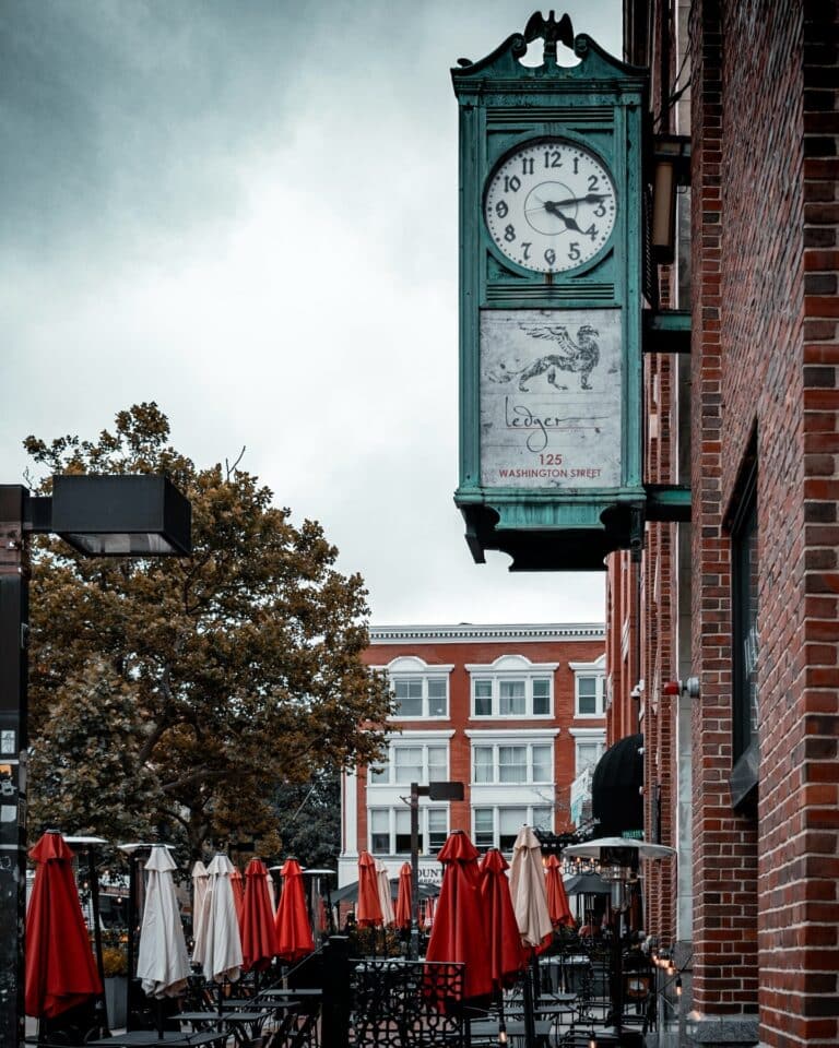 A historic clock hanging above the entrance of Ledger Restaurant in Salem, Massachusetts, with colorful umbrellas and a bustling outdoor dining area below.
