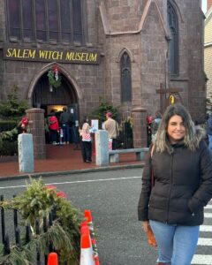 A woman in a black winter coat stands in front of the Salem Witch Museum in Salem, Massachusetts, with holiday decorations, wreaths, and people entering the museum.