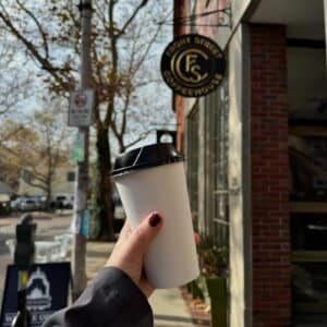 A hand holding a coffee cup in front of Front Street Coffeehouse in Salem, Massachusetts on a sunny day. The café’s sign hangs above the entrance, and fall trees line the street.