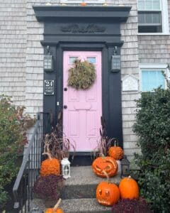 A pastel pink door in Salem, Massachusetts, framed by a black doorway and cedar shingles. The entrance is decorated with carved jack-o'-lanterns, bumpy pumpkins, autumn plants, and a natural wreath, creating a charming Halloween vibe.