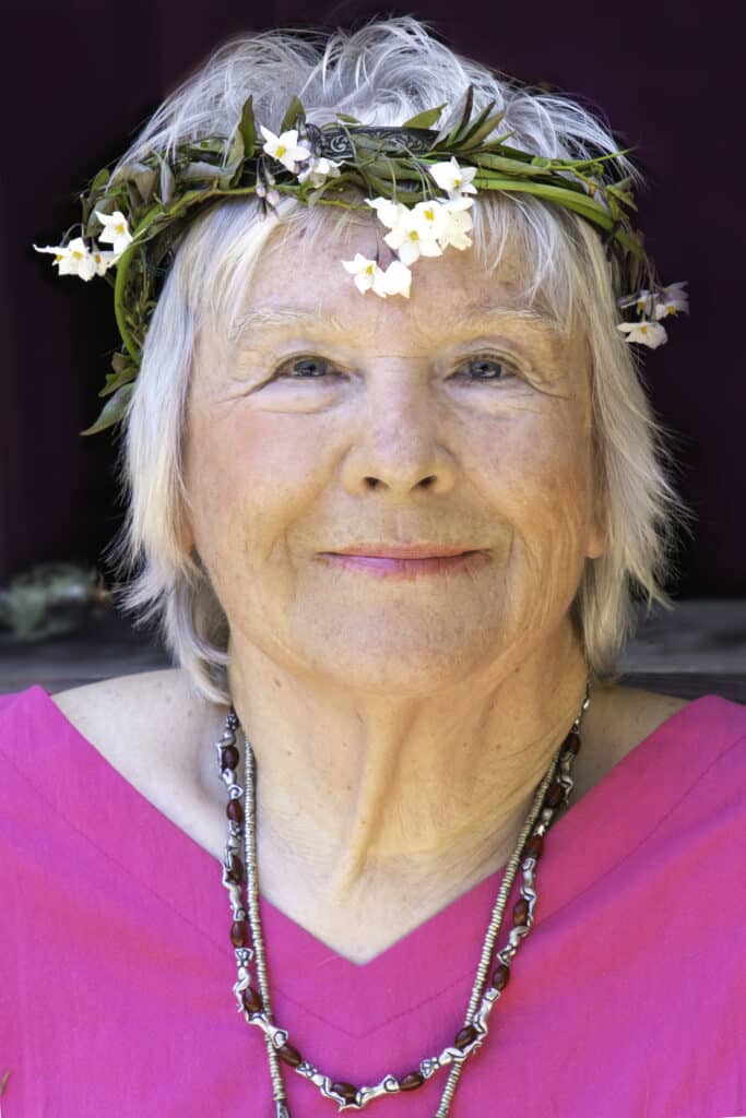 Zsuzsanna Budapest, a renowned feminist spiritual leader and founder of Dianic Wicca, is depicted in this image. She wears a pink outfit and a floral crown made of delicate white flowers and green leaves, symbolizing her connection to nature and the divine feminine. Her silver hair frames her serene expression, embodying wisdom and empowerment. Adorned with layered necklaces, she exudes a graceful presence that reflects her role as a trailblazer in modern feminist spirituality. The warm lighting complements her aura of strength and nurturing energy.