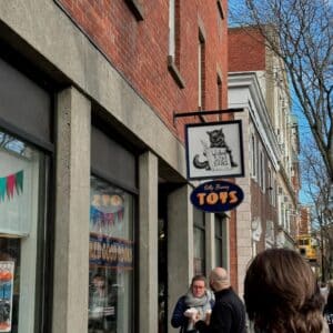 A street view of Wicked Good Books and Silly Bunny Toys in Salem, Massachusetts, shows their shop signs hanging from a red brick building. The bookstore sign features a cat reading a book, and the toy store sign has bright, playful lettering. A few people are walking and chatting along the sidewalk.