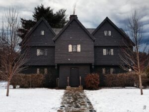 Snow-covered Witch House in Salem, Massachusetts, with bare trees and smoke rising from the chimney on a cloudy winter day.
