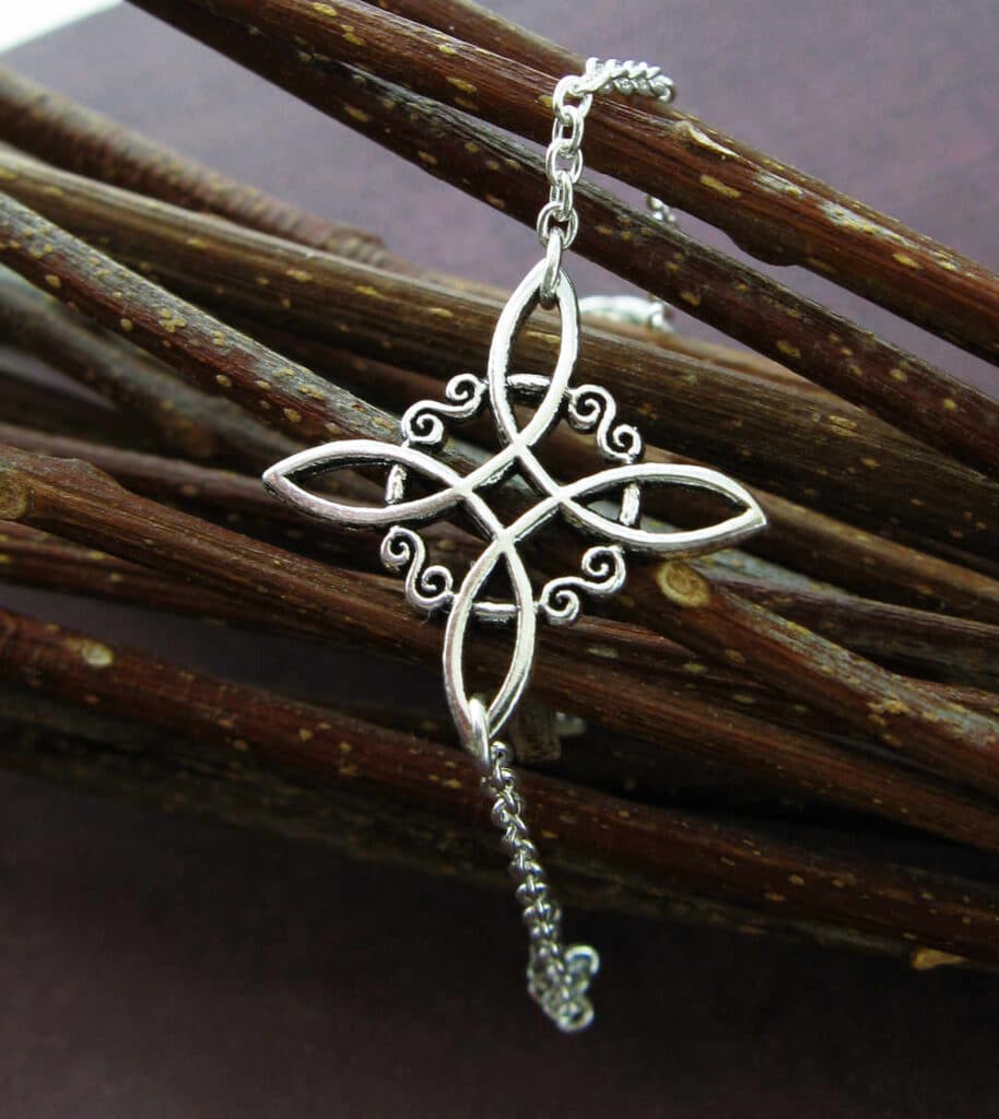 A silver Witch’s Knot pendant with intricate interwoven loops and delicate scrollwork, resting against a bundle of twigs. This mystical symbol is associated with protection, binding energy, and magical intention in Wiccan and pagan traditions. The polished metal contrasts with the natural background, emphasizing its role as a talisman for shielding against negative forces and enhancing spiritual energy.